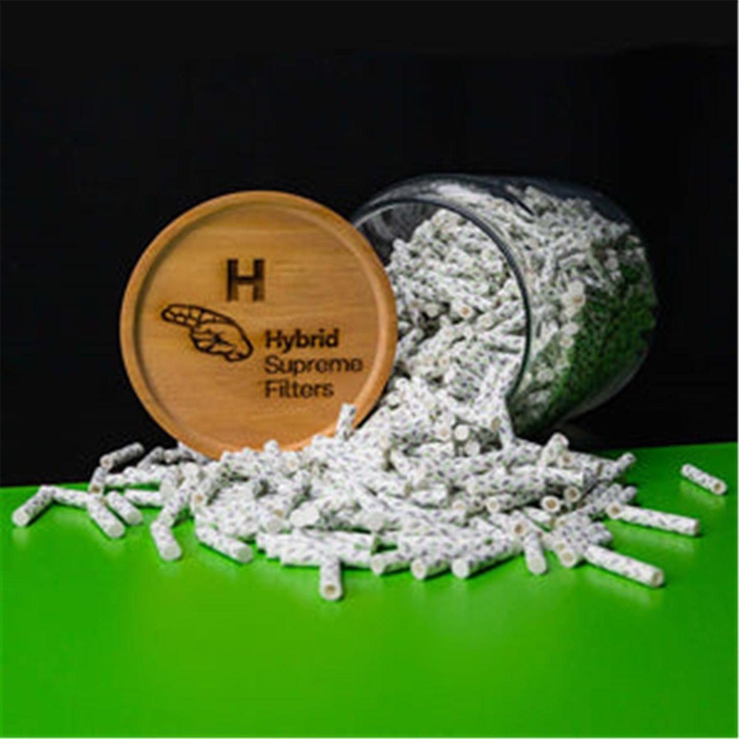 Hybrid Supreme Filters 1000er Glasdose – geöffnetes zylindrisches Glasgefäß mit Holzdeckel und eingeprägtem Hybrid-Logo, aus dem zahlreiche weiß-graue Filter auf grüne Fläche herausgefallen sind, schwarzer Hintergrund